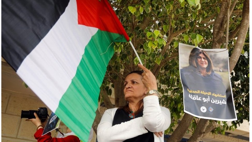 سيدة ترفع علم فلسطين بجوار صورة لشيرين أبو عاقلة.
